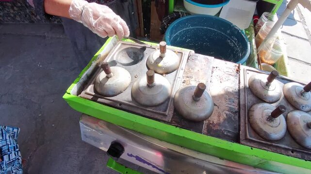 Video of Hand places lid on Indonesian traditional pancake maker. Food vendor prepares grilled pancakes. Bright green appliance sits on street corner. Steam rises gently from cooking units. 4K Footage