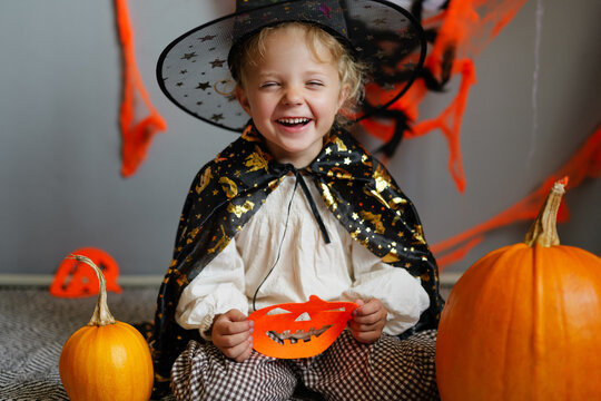 A child is sitting among pumpkins wearing a costume with a cape and a witch hat. The child holds a mask and shows happiness in a playful setting