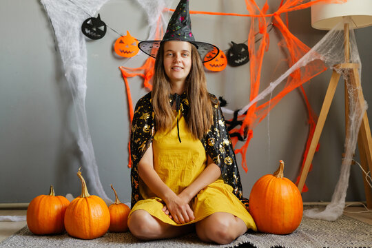 A child sits on the floor surrounded by pumpkins wearing a witch costume. Halloween decorations hang on the wall, creating a festive atmosphere