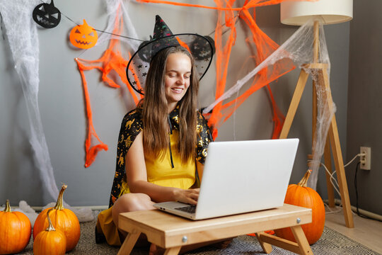 A girl wearing a witch costume sits on the floor and types on a laptop. The room has Halloween decorations such as pumpkins and cobwebs