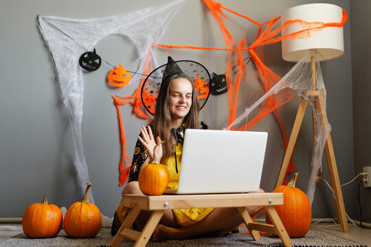 A woman dressed in a witch costume waves at her computer during a Halloween celebration at her house. Pumpkins and decorations set the scene for the event