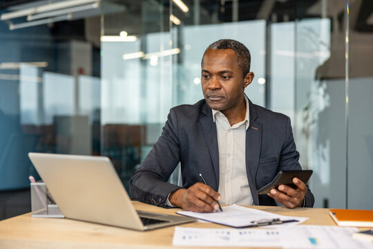 Businessman analyzing financial data, calculating figures, and writing notes on documents at his desk, demonstrating dedication to corporate finance and accounting tasks