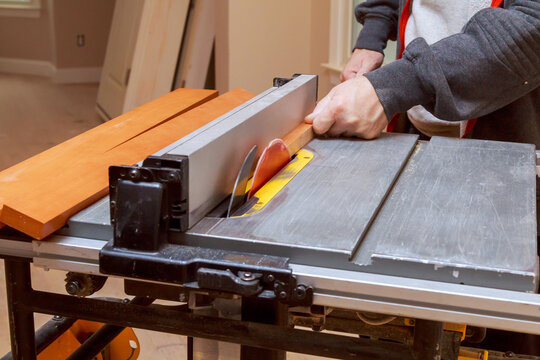 Worker stands at table saw, cutting piece of wood various wooden boards are