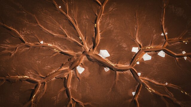 Aerial view of parched riverbed with intricate network of dry cracks, interspersed with white salt deposits and glistening gold flakes.
