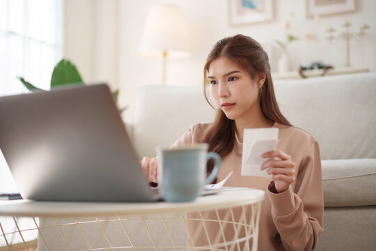 Asian Woman reviewing receipts and managing household budget with laptop at home, supporting financial planning and expense control.