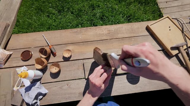 First-person POV of applying brown protective wood stain to a handcrafted wooden bird feeder component with a brush. 