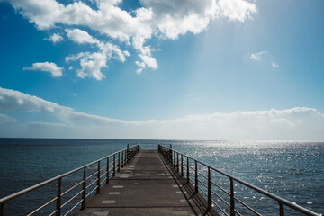 Fototapeta premium A peaceful ocean pier stretching toward the horizon, with sparkling water, dramatic clouds and bright sunlight creating a calm coastal scene.