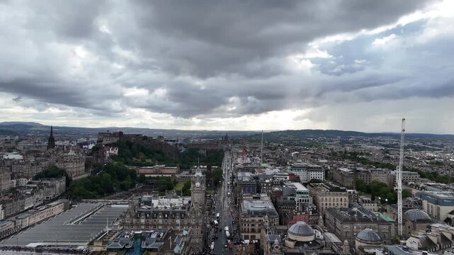 Wide panoramic aerial view of Edinburgh cityscape under dramatic cloudy sky, featuring Edinburgh Castle, Balmoral clock tower, Waverley Station, and Princes Street, 4k drone footage