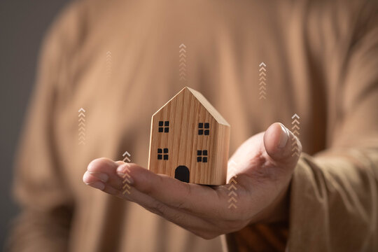 Real estate appreciation concept with a wooden house model held in hand and rising arrows symbolizing increasing property values, housing market growth, home investment, equity gains, wealth building.