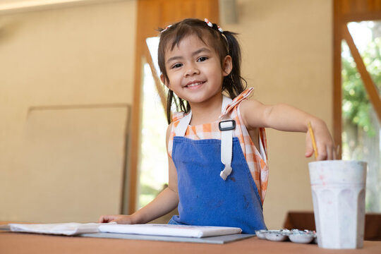 Happy Asian little girl painting on canvas in a art classroom, wearing an apron and smiling with confidence. Creative learning, childhood education, imagination, artistic development and fun activity.