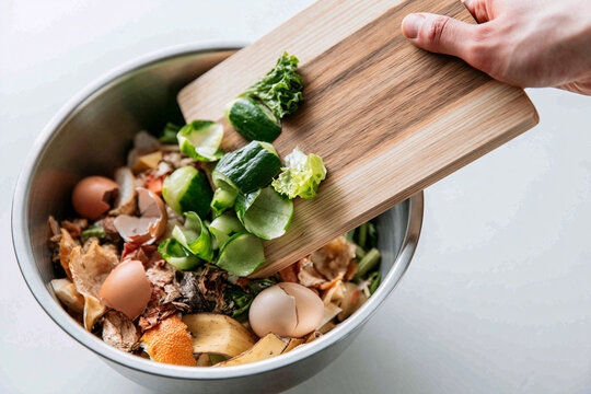 Top-down shot of a person's hand holding a cutting board over a stainless steel bowl filled with organic kitchen food scraps for composting.