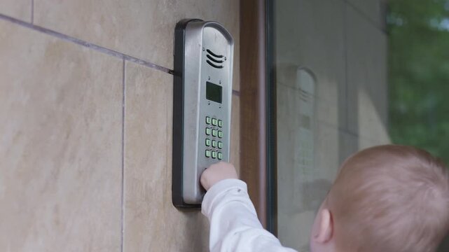 Young child presses buttons on a modern intercom system mounted on a wall, with a glass door reflecting the surrounding greenery in the background