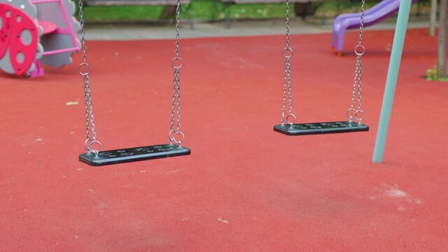 Two empty swings in a playground with red rubber flooring, surrounded by colorful play equipment and benches, indicating a serene outdoor environment for children