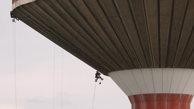 Suspended rope access technicians perform maintenance under a massive concrete water tower. This extreme industrial job highlights professional safety and courageous infrastructure repair.