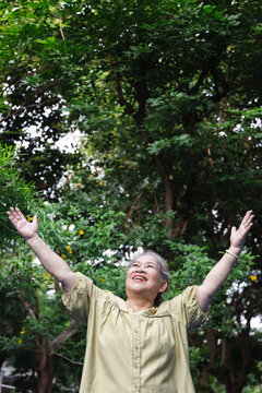 Happy senior asian woman breathing fresh clean air in urban park during summer wellness sunlight exposure, front view
