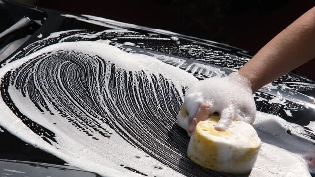 Close-up of a hand washing a black car with a soapy sponge, creating rich foam during vehicle cleaning, showcasing automotive care, maintenance, and detailing