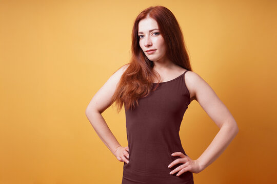 Confident young woman with long red hair posing with hands on hips in brown strappy dress on yellow studio background