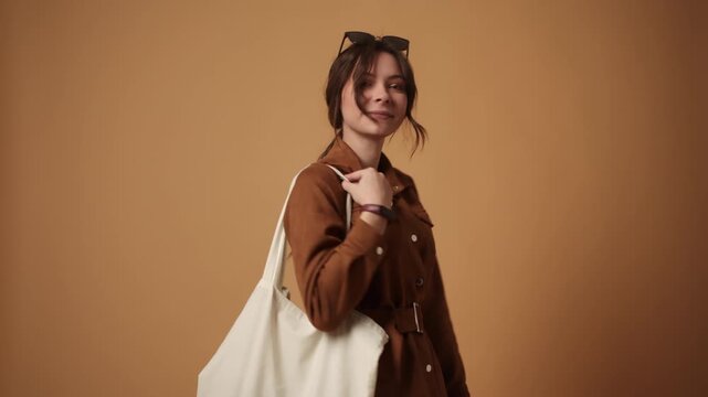 Pretty young woman posing with a white tote bag in a studio with a warm brown background and smiling at the camera