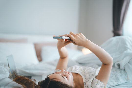 Happy smile asian woman waking up in bedroom using smartphone internet app relax on bed