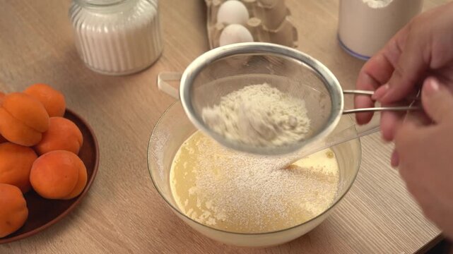 Pastry chef woman sieves flour into egg batter in a glass bowl while preparing dough for homemade baking.