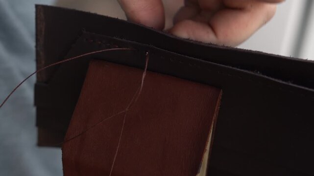 Leather craftsman hand sewing a handmade wallet with needle and thread in a workshop, close up of the artisan creating leather goods