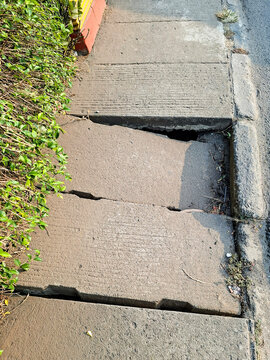 A damaged concrete sidewalk with broken slabs, visible cracks, and uneven surfaces next to green bushes along a city street.