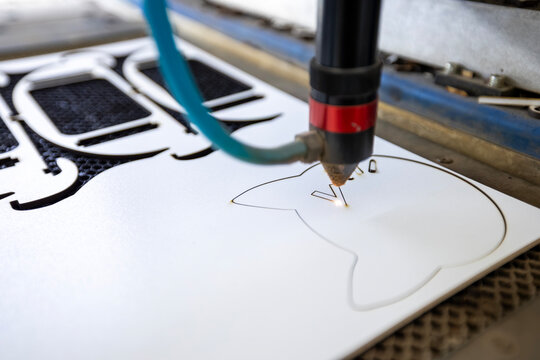 Laser cutting machine engraving custom patterns on white material with precision beam nozzle inside industrial workshop, bright focused glow marks surface detail, creating intricate shapes during