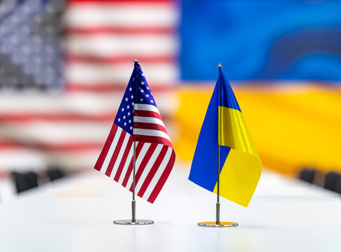 Miniature national flags displayed on a white conference table with blurred country banners in the background representing international diplomacy and cooperation between two nations during a formal