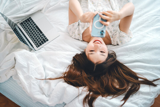 Happy smile asian woman waking up in bedroom using smartphone internet app relax on bed