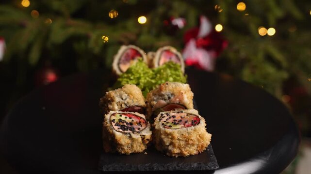 Christmas themed tempura sushi rolls with tobiko caviar and salmon on a black slate platter with a festive decorated tree and sparkling bokeh lights in the background