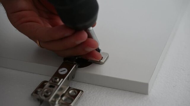Close-up video of a technician installing and adjusting cabinet door hinges using a screwdriver and power drill. Furniture assembly, repair, and maintenance concept
