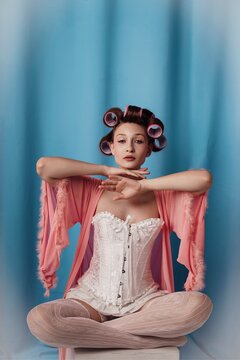 Sitting elegantly in a pastel hue, a model embraces vintage glamor with hair rollers and a flowing robe