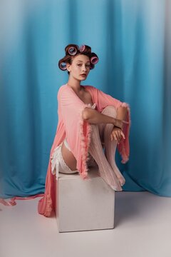Sitting elegantly in a pastel hue, a model embraces vintage glamor with hair rollers and a flowing robe