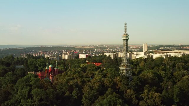 Petrin Lookout Tower rises above green trees beside Saint Lawrence Church in Prague. Historic landmark stands on hill above city background. European travel destination offers rich culture.