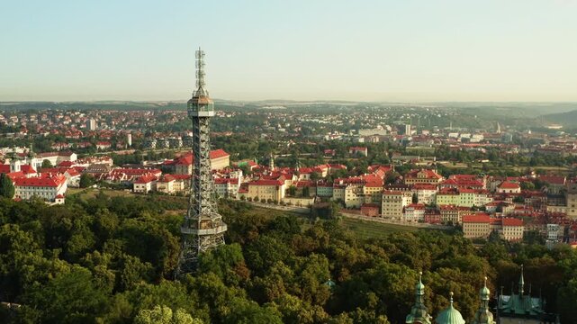 Petrin Lookout Tower stands above green forest on Petrin Hill in Prague. Historic city panorama with red tile roofs stretches under sunny sky. European capital landmark for travel concept.