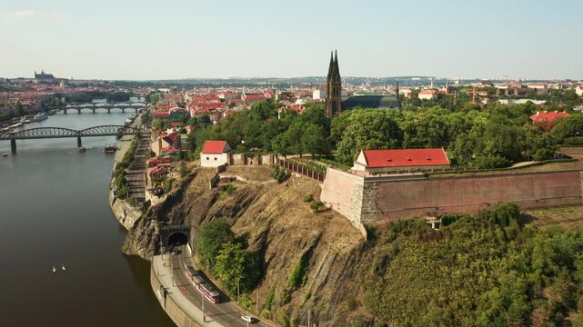 Vysehrad fortress and Basilica of St. Peter and Paul stand on a cliff in Prague. A tram enters a road tunnel below historic brick walls along the Vltava riverfront in European capital city.