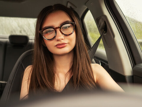 Young woman with massive glasses drives modern car. Confident and calm driver with small experience. Car interior shows clean, sleek design. Outside view shows green landscape.