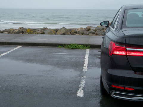 Black luxury car is parked in a car park with stunning view on the ocean in a cold morning mist. Travel and tourism. Sky is overcast and pale. Vehicle rear lights glow red.