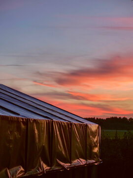 Sunset colors glow behind greenhouse roof. Sky shows orange, pink, and blue hues. Greenhouse structure stands in open field. Trees line distant horizon. Calm evening atmosphere captured
