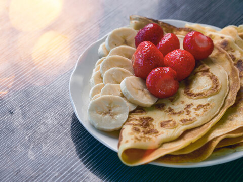 Stack of golden-brown pancakes on white plate. Fresh strawberries and sliced bananas beside pancakes. Soft and dreamy look, Hand made simple country meal. Dark wooden surface background