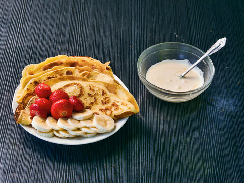 Stack of golden-brown pancakes on white plate. Fresh strawberries and sliced bananas beside pancakes. Small glass bowl with creamy sour cream and spoon. Dark wooden surface background