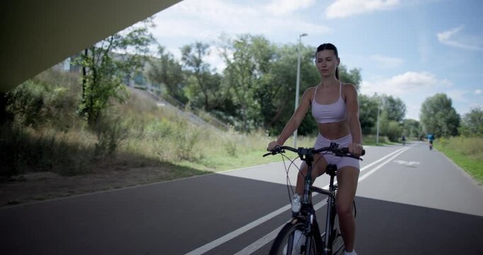 Young beautiful athletic woman in sportswear walking and riding a bicycle in the city in an urban park with a bridge and benches