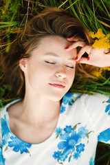 Naklejka premium Beautiful smiling cute blonde young woman lies in green grass with eyes closed, red nails resting on her forehead, wearing a floral dress and enjoying a peaceful summer moment outdoors