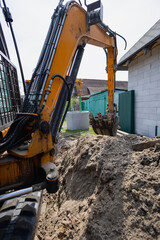 Fototapeta premium A digger works in a backyard, creating a trench for construction. Soft earth is piled high, and nearby structures are visible under a clear sky