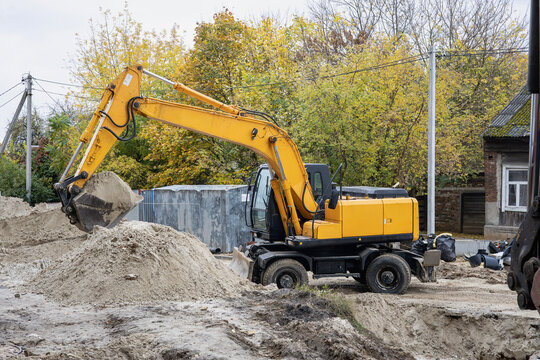 A yellow excavator is digging dirt in a construction site. Trees with autumn leaves are in the background. The site has piles of sand and equipment