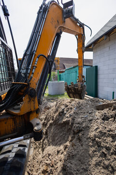 A digger works in a backyard, creating a trench for construction. Soft earth is piled high, and nearby structures are visible under a clear sky
