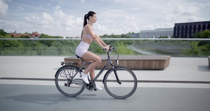Young beautiful athletic woman in sportswear walking and riding a bicycle in the city in an urban park with a bridge and benches
