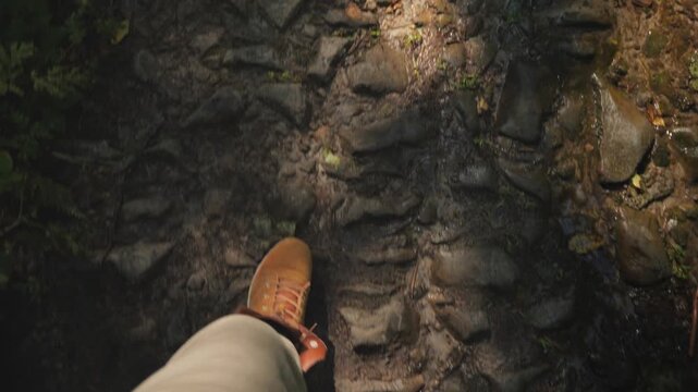 Pov of person wearing brown leather boots walking on rocky forest trail along small stream.