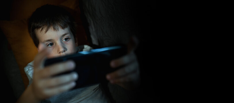 A 6-7-year-old boy plays on his mobile phone and checks social media while lying down instead of sleeping. Digital addiction in children, banner copy space