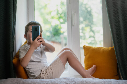 A 6-7 year old child sits on a windowsill at home with a mobile phone and plays. Mental health issues, unlimited gadgets, FOMO, harm to the child's psyche.